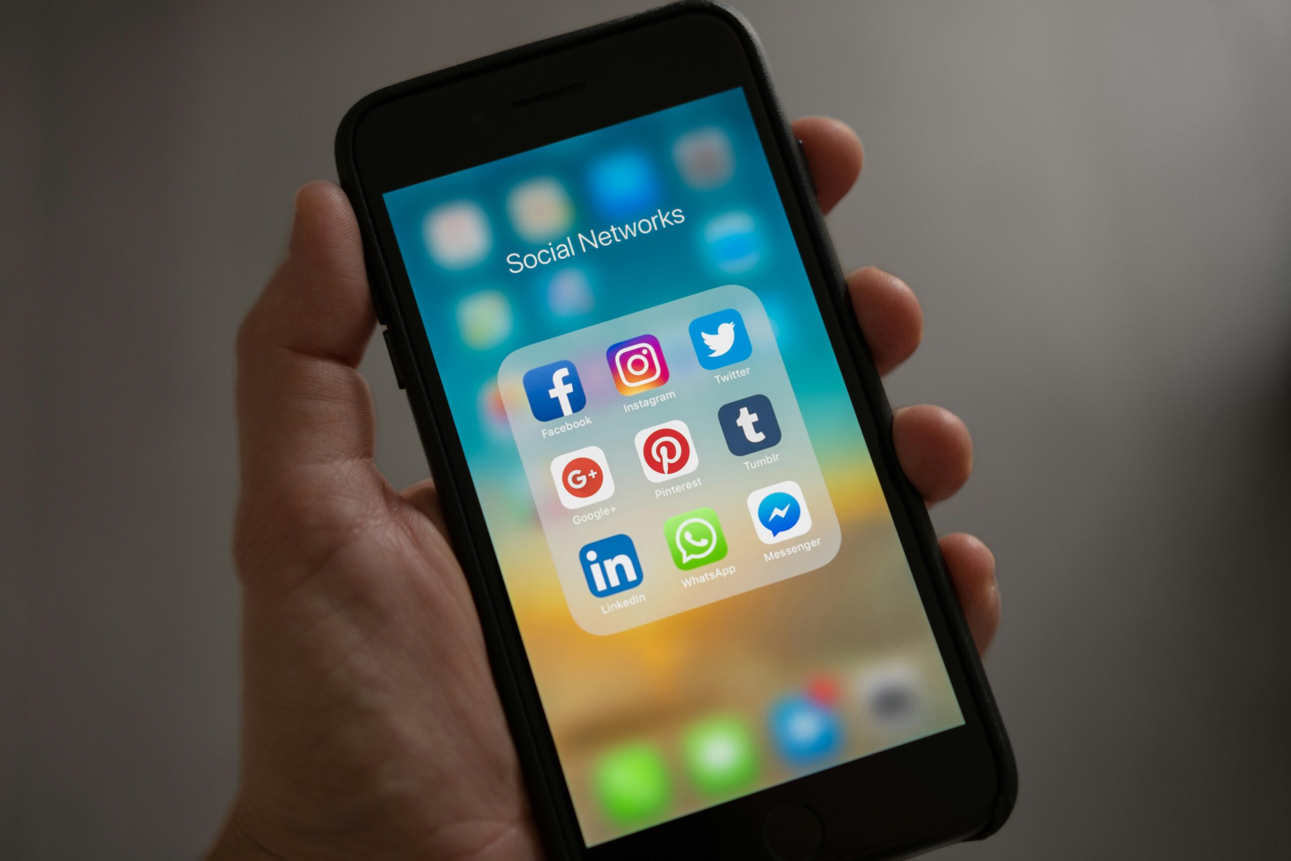 A hand holding a smartphone displaying a folder labeled "Social Networks" with various app icons: Facebook, Instagram, Twitter, Google+, Pinterest, Tumblr, LinkedIn, WhatsApp, Messenger.